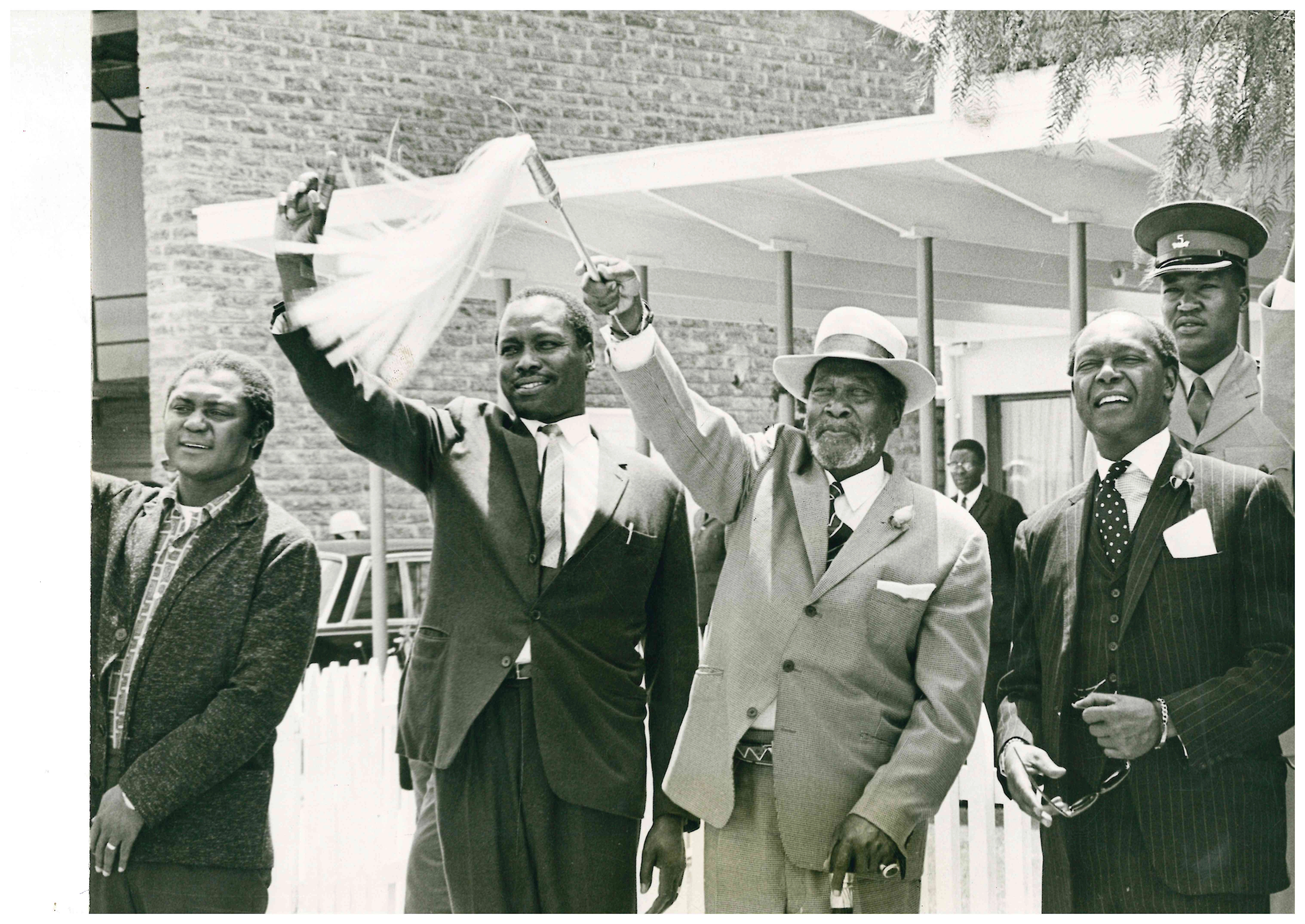 President Jomo Kenyatta dressed in a light-coloured suit, patterned tie and brimmed hat, waving his ceremonial fly whisk while his Vice President Daniel Moi stands to his right waving the pointing finger (the hand sign for their Kanu party). To their right is Minister for Economic Planning Tom Mboya and to the left Attorney General Charles Njonjo.