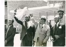 President Jomo Kenyatta dressed in a light-coloured suit, patterned tie and brimmed hat, waving his ceremonial fly whisk while his Vice President Daniel Moi stands to his right waving the pointing finger (the hand sign for their Kanu party). To their right is Minister for Economic Planning Tom Mboya and to the left Attorney General Charles Njonjo.